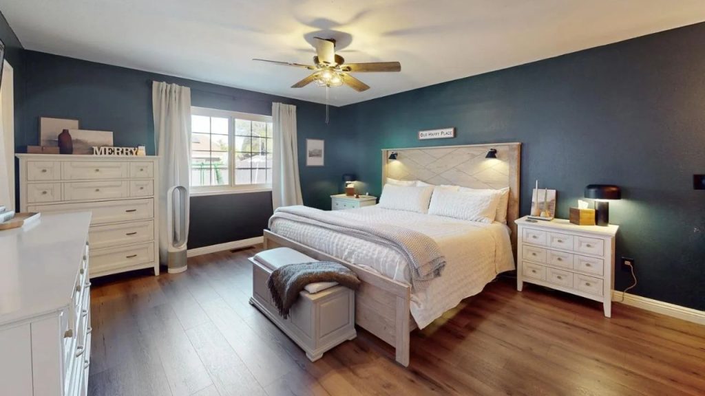 A cozy master bedroom with a teal wall, white bed, matching dressers, ceiling fan, and soft-curtained window.