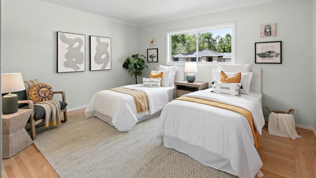 A cheerful guest bedroom with two beds, yellow throws, and a sunny window overlooking trees.