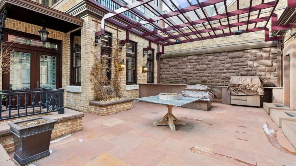 An intimate courtyard with a stone-tiled patio, pergola canopy, wooden table, and brick walls, featuring classic lanterns and a covered outdoor kitchen area.