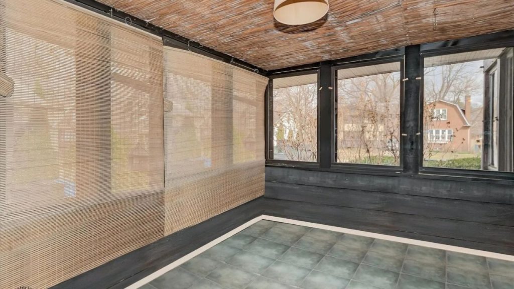 A rustic sunroom with large windows, bamboo shades, dark wooden walls, a woven mat ceiling, a pendant light, and tiled floors, offering a cozy outdoor view.