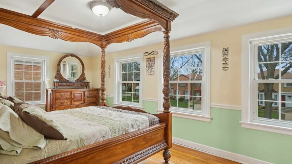 Bright bedroom with carved canopy bed, green accent walls, and large windows.