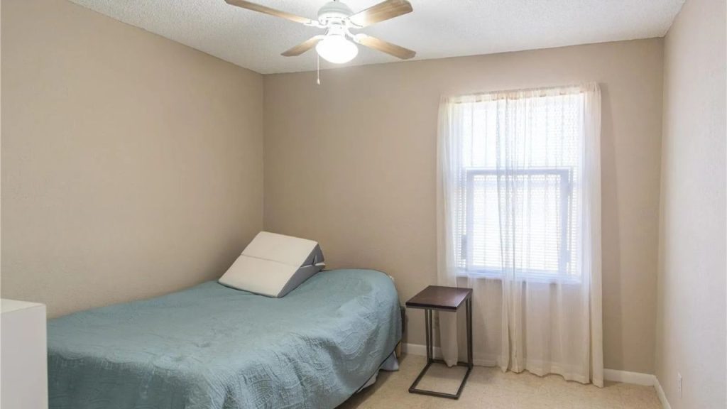 A minimalist bedroom with beige walls, a blue quilted bed, ceiling fan, and sheer curtains.