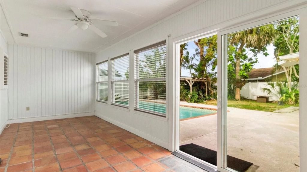 A bright porch with white shiplap walls, terracotta tiles, large windows with shutters, a ceiling fan, and a view of a pool and lush backyard.