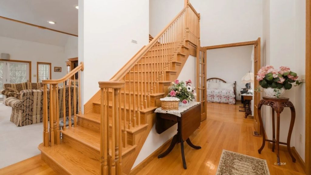 A grand wooden staircase with warm tones, leading to a cozy bedroom and a spacious living area.