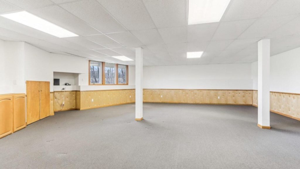 A spacious basement with gray carpet, wooden wainscoting, white columns, and high windows, offering a bright and versatile vibe.