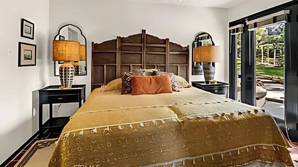 A cozy bedroom with a wooden headboard, golden bedspread, arched mirrors, and glass doors opening to a green view.