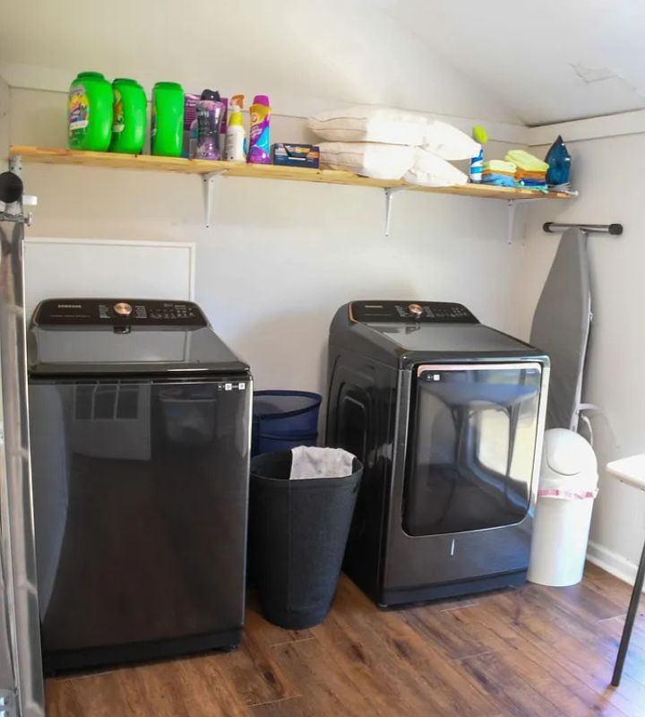 A compact laundry room with black washers, dryers, a wooden shelf, and ironing board on hardwood floors.