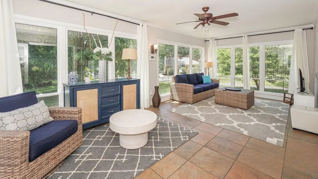 A sunlit family room with woven blue furniture, large windows, and a stylish cabinet under a ceiling fan.
