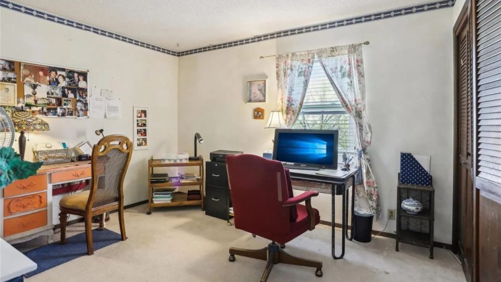 A cozy home office with an orange desk, red chair, floral curtains, and photo board, featuring a monitor and wooden shutters.
