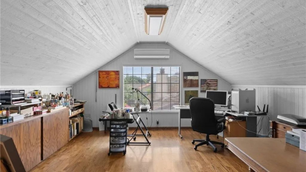 A quiet attic workspace with hardwood floors, a desk, computer, art supplies, and large window.