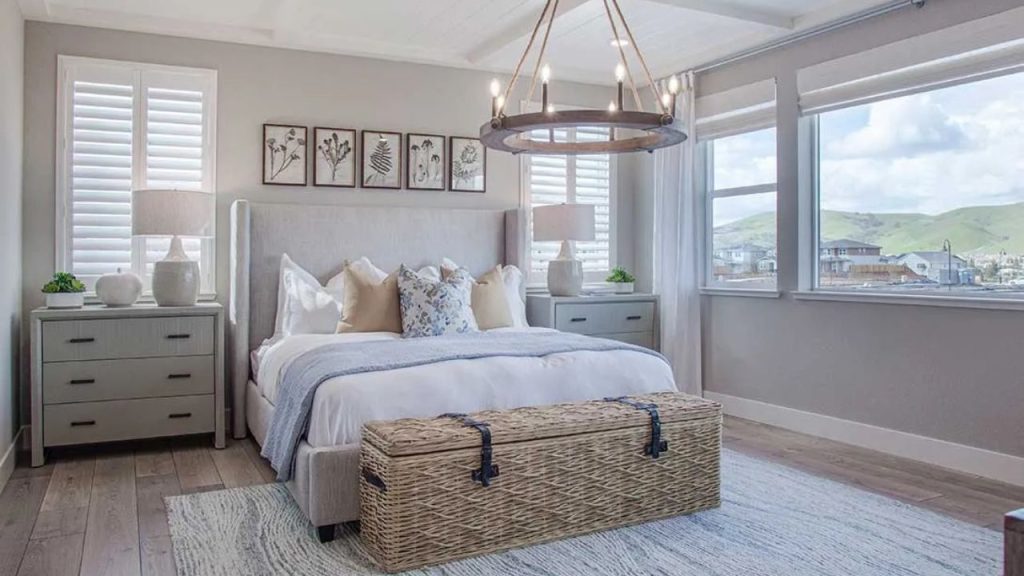 An elegant bedroom with a gray bed, white linens, patterned pillows, botanical wall art, a woven chandelier, large windows with shutters, and a wicker storage bench on a light wood floor.