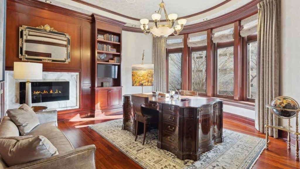 A refined home office with a mahogany desk, chandelier, built-in bookshelves, a modern fireplace, and large windows with cream curtains, set against hardwood floors.