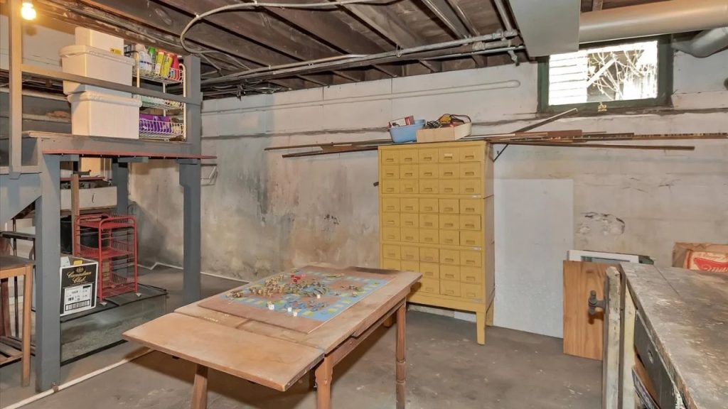 An eclectic basement workshop with exposed pipes, a wooden table with a board game, a yellow vintage cabinet, metal shelves with bins, and a small window letting in light.