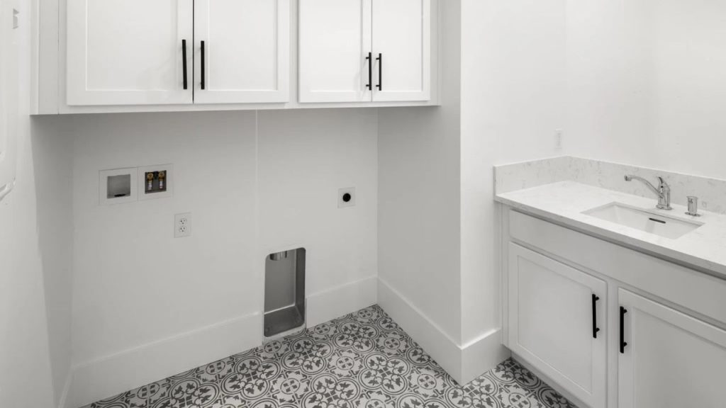 A laundry room with white cabinets, a marble countertop with a sink, patterned gray tile flooring, and a small pet door on the wall.