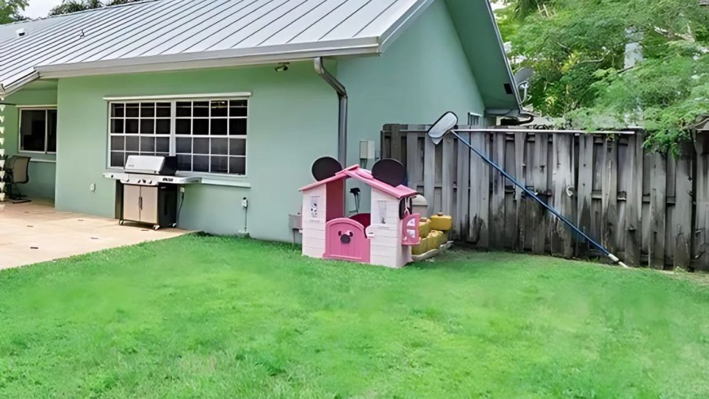 A green backyard with a pink playhouse, grill, and wooden fence.