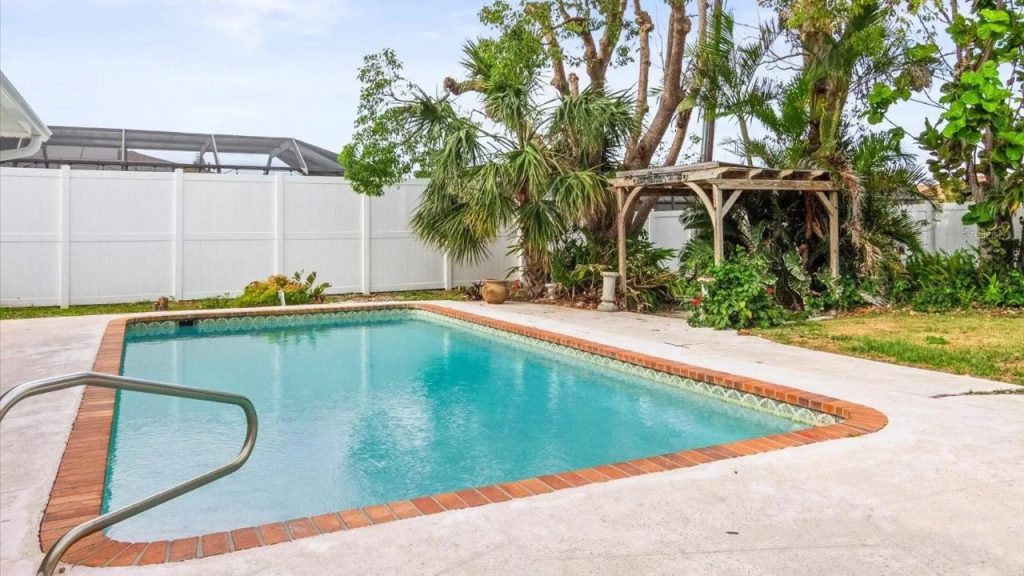A tropical backyard with a sparkling pool, brick edge, handrail, lush greenery, a rustic pergola, and a white fence under a sunny sky.