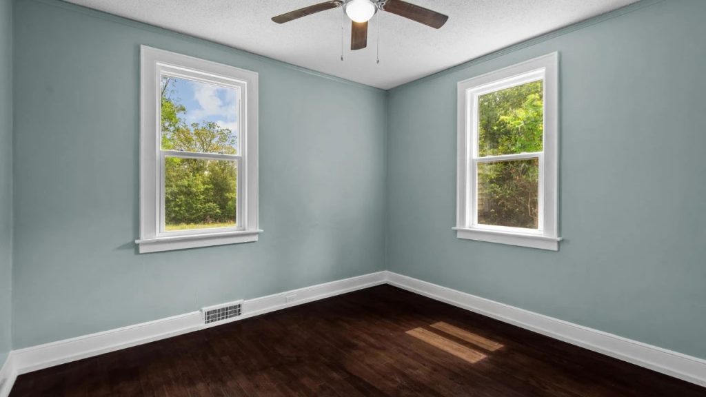Peaceful bedroom with green walls, dark floors, and two windows overlooking greenery.