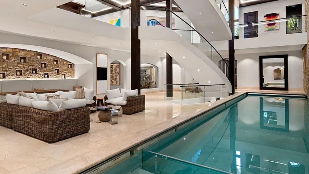 An indoor pool with glass edge, wicker seating, and a curved staircase leading to an art-filled upper level.