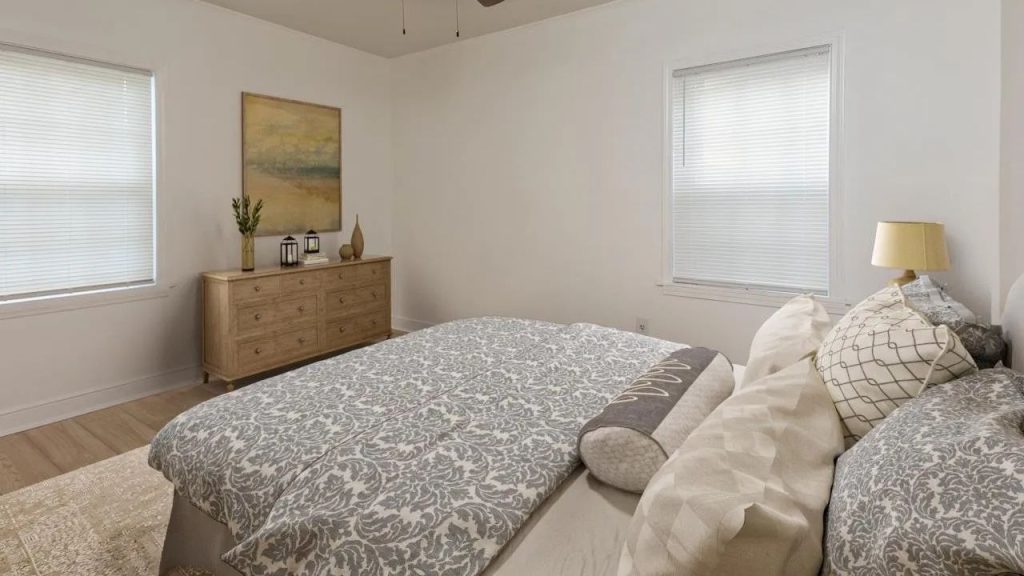 A cozy bedroom with a patterned bed, wooden dresser, lamp, and large windows with blinds.