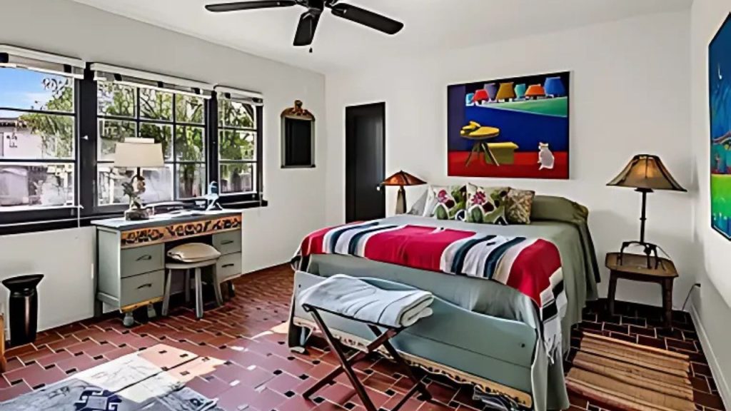 A cheerful bedroom with a red and blue striped bed, terracotta tiles, large windows, and vibrant artwork.