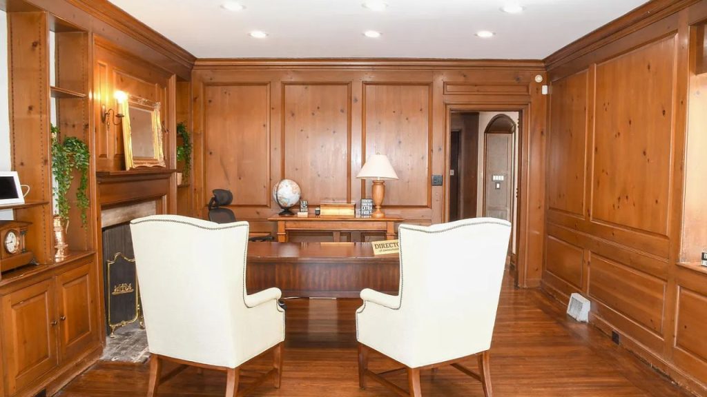 A classic study with wooden paneling, a desk, white armchairs, and a fireplace.