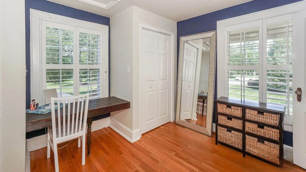 A quiet home office with a blue accent wall, wooden desk, large windows, and a woven basket dresser.