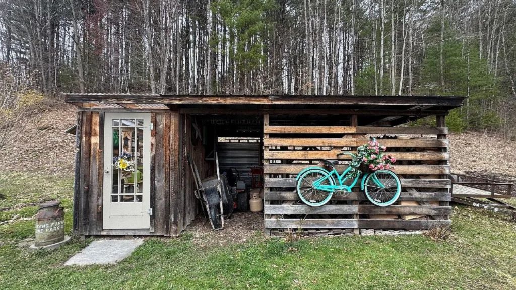 A rustic shed with a turquoise bike, floral wreath, and wheelbarrows, set against a wooded hillside.