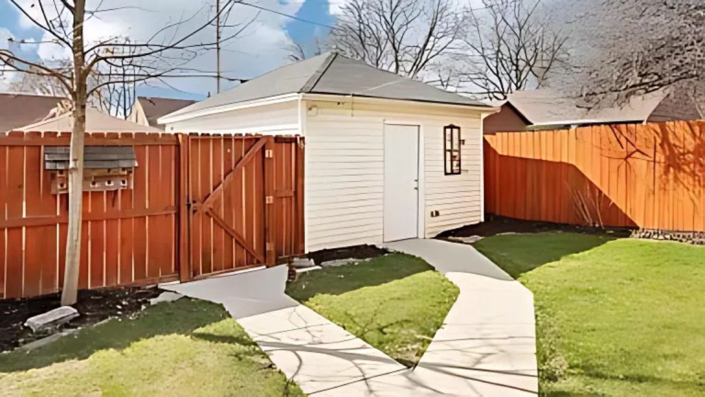 A cozy white shed with a small window, set against an orange fence in a green backyard.