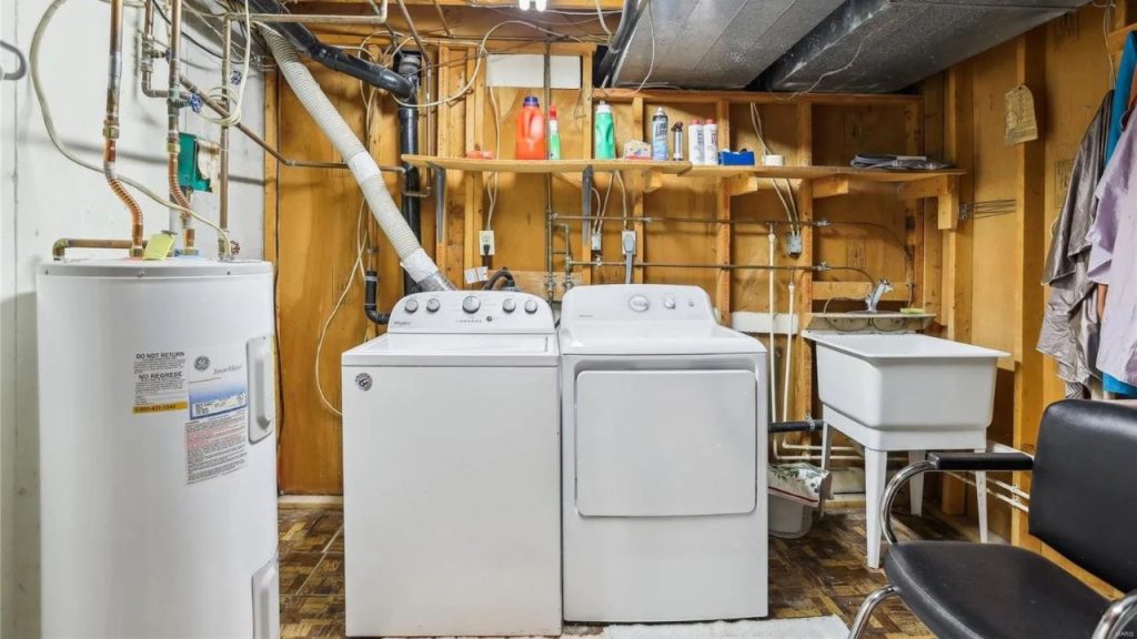A rustic laundry room with white washer, dryer, utility sink, and wooden shelves, featuring exposed pipes and a black chair.