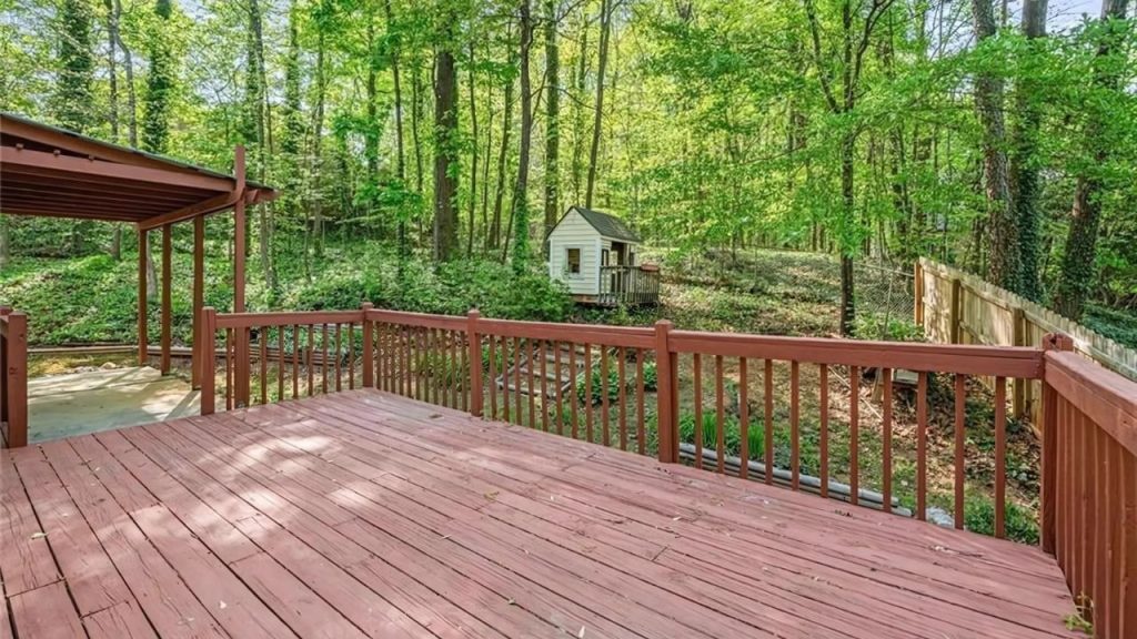 A wooden deck with a shaded roof, overlooking a forest with a playhouse and fenced backyard.