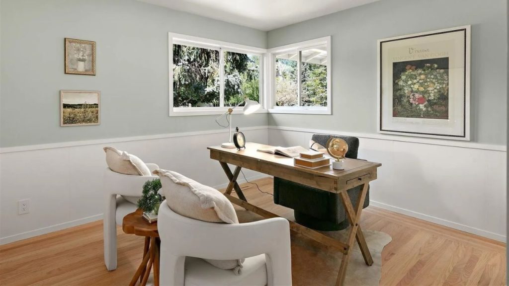 A cozy home office with a wooden desk, white chairs, and large windows overlooking greenery.