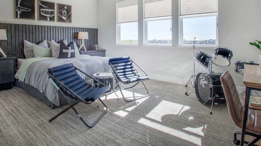 A modern bedroom with a dark headboard, gray bed with patterned pillows, blue lounge chairs, a drum set, music-themed wall art, large windows with shades, and a light gray carpet.