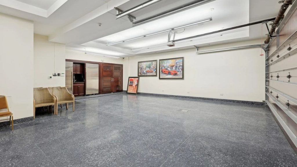 A spacious garage with a polished concrete floor, built-in refrigerator, wicker chairs, and vintage car artwork on the walls, lit by bright overhead lights.