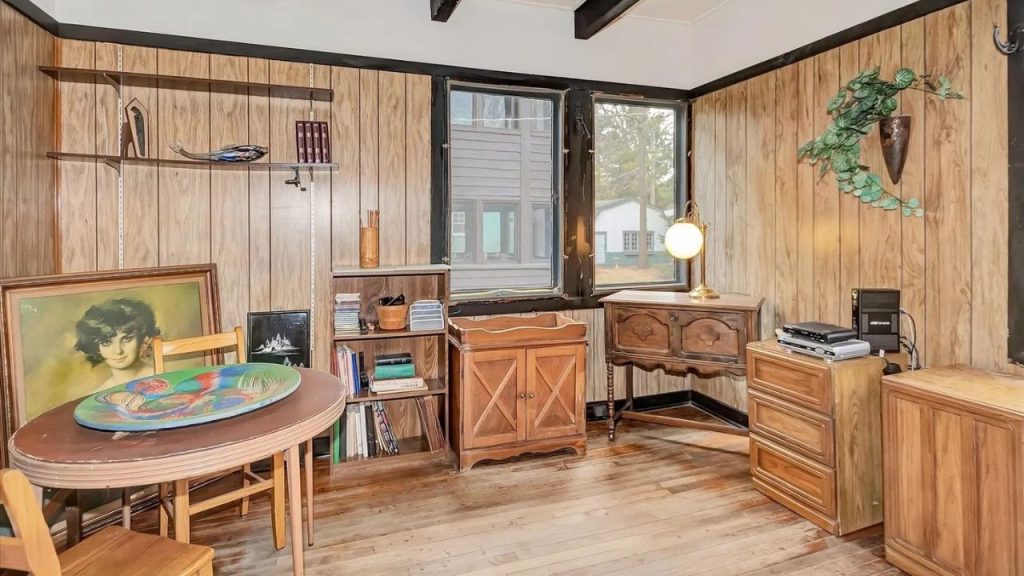 A nostalgic den with wooden paneling, a round table with abstract art, a bookshelf with books, a carved cabinet, large windows, and a cascading plant on the wall.