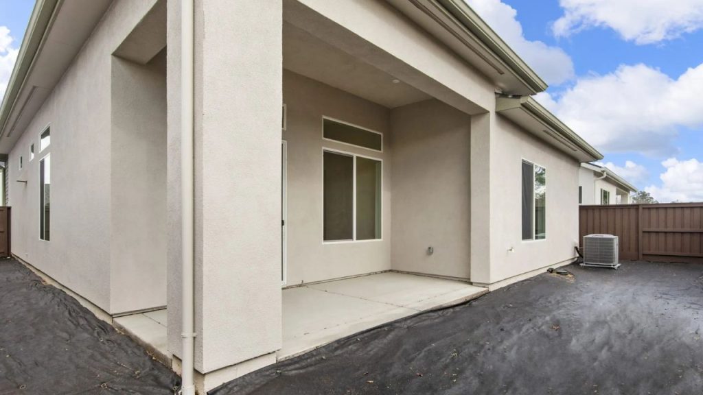 A covered back patio with a concrete slab, neutral exterior walls, large windows, and a wooden fence under a partly cloudy sky.