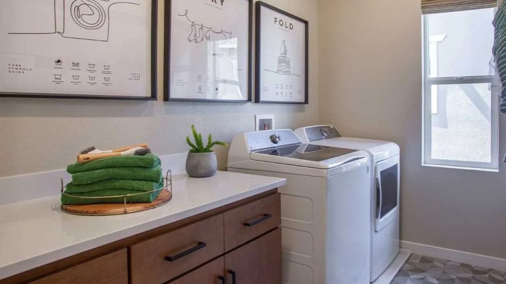 A bright laundry room with white washer and dryer, wooden cabinets, green towels, and playful wall art.