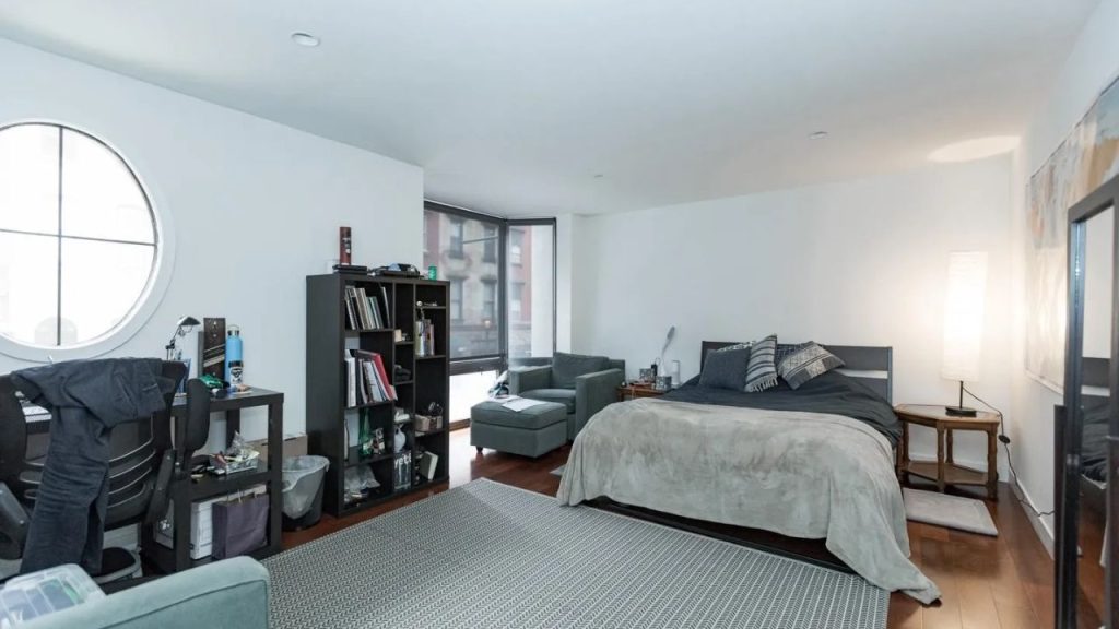 Studio bedroom with bed, desk, bookshelves, and large round window.