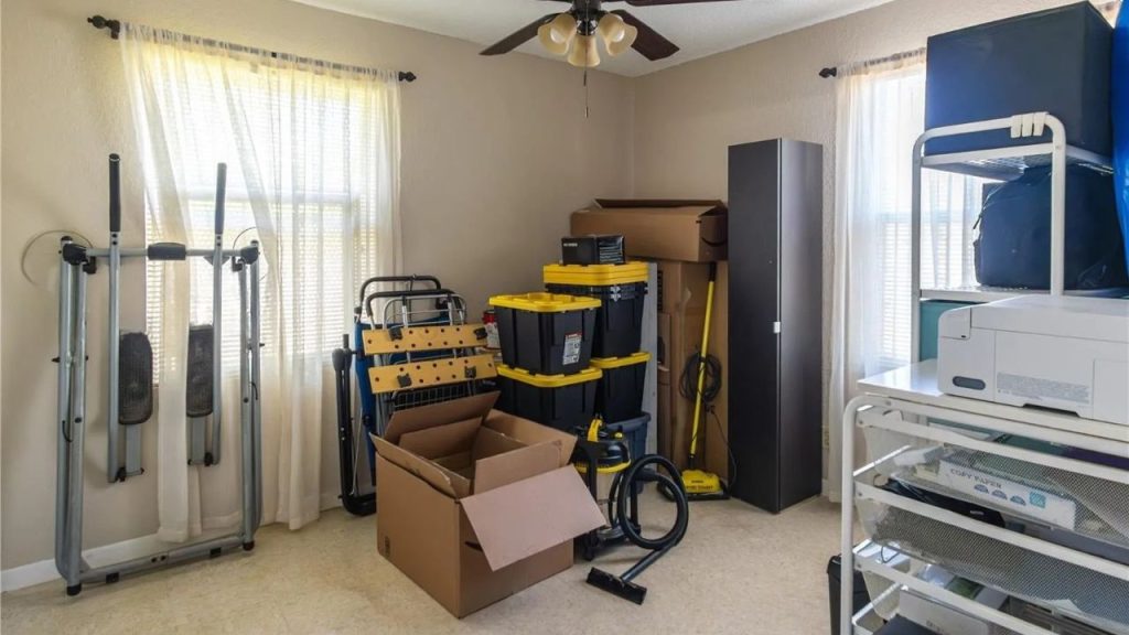 A storage room with bins, workout gear, a vacuum, printer, and ceiling fan under sheer curtains.