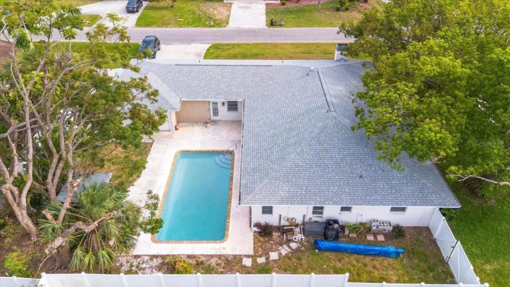 A single-story suburban home with a light gray exterior, shingle roof, white garage door, pale blue shutters, and a white fence surrounded by lush trees.