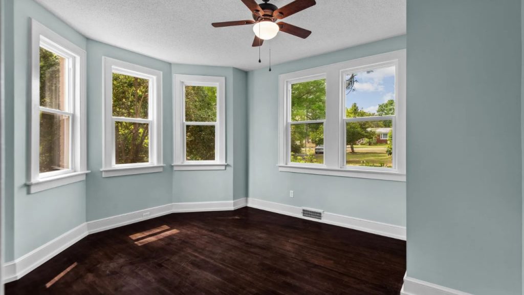 Bright bay window room with green walls, dark wood floor, and tree-filled views.