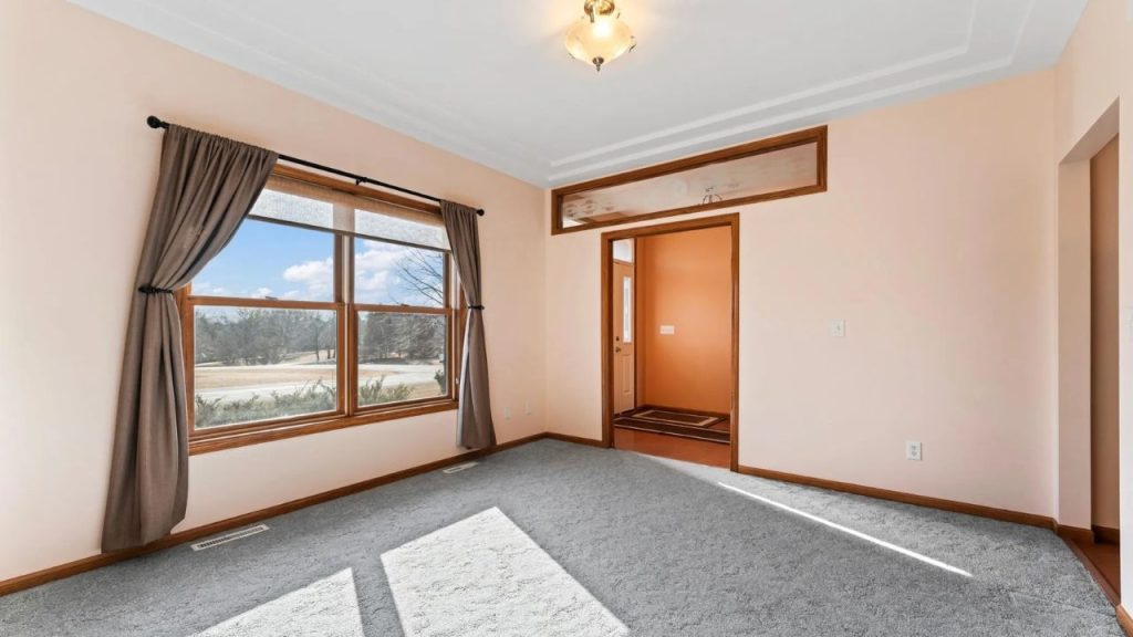 Bright front room with peach walls, large window, and gray carpet.