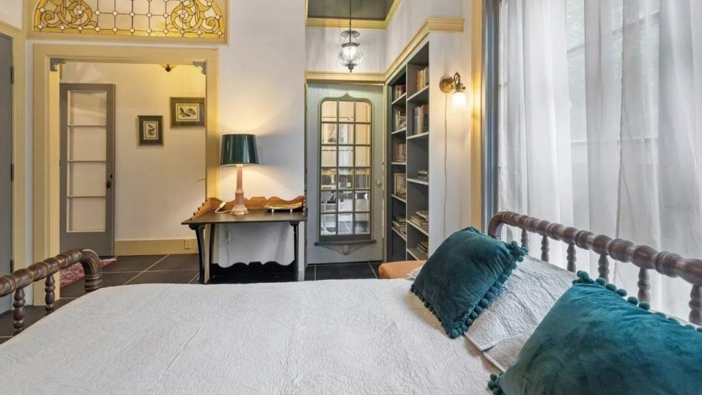 A cozy bedroom with a wooden bed, teal pillows, a desk with a green lamp, built-in bookshelves, and arched doorway with stained glass.