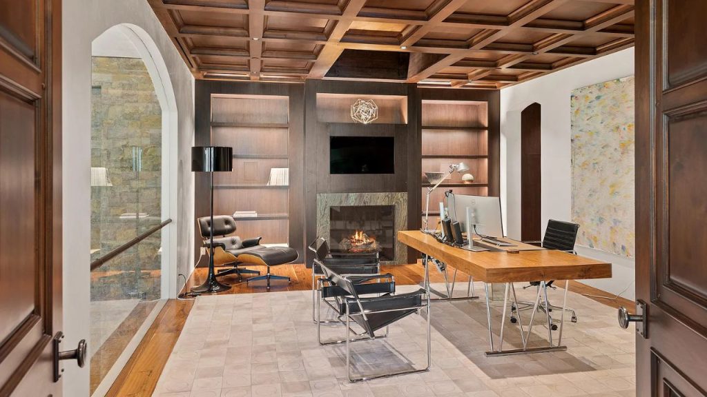 A stylish office with a wooden desk, black chairs, a fireplace, and an Eames lounge chair, featuring a coffered ceiling and abstract art.