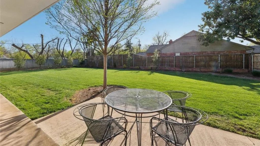 Large grassy backyard with patio and outdoor seating under a tree.