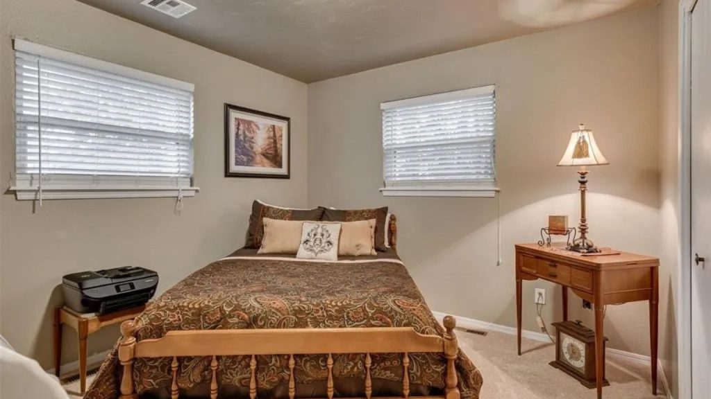 Cozy guest room with a wooden bed, bedside lamp, and windows, featuring soft carpet and a small table with a printer.