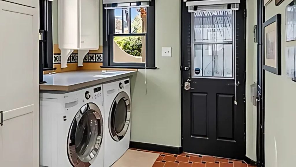 A cozy laundry nook with two white washers, terracotta tiles, a yellow backsplash, and a black door.