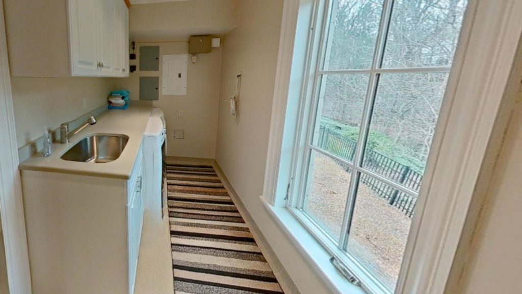 A cozy laundry room with a sink, washer, white cabinets, and a large window overlooking greenery.