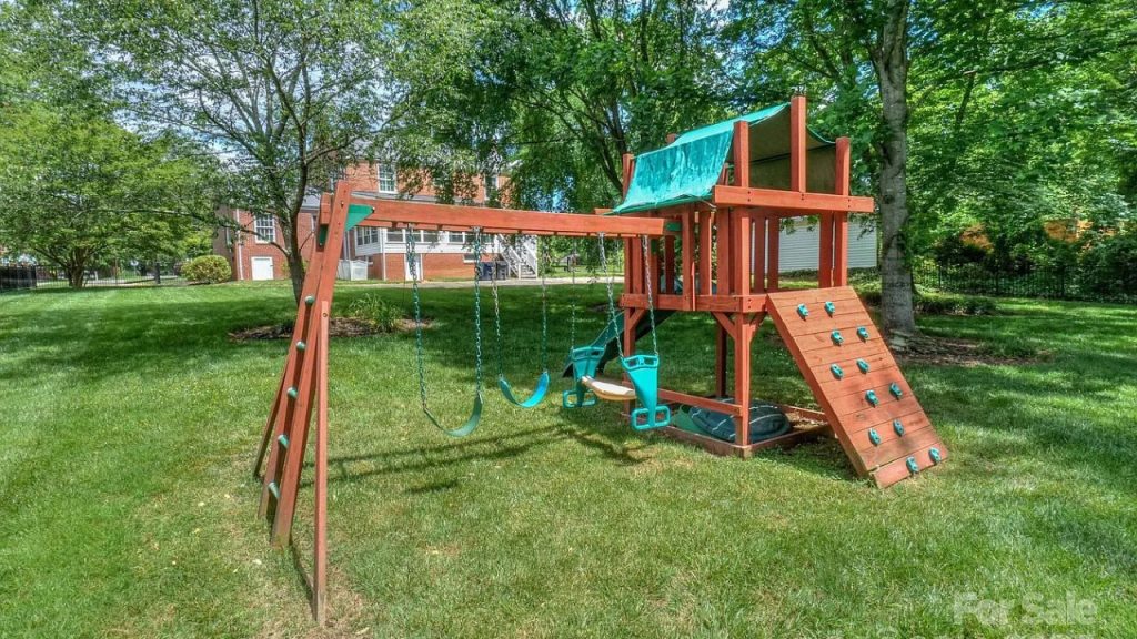 A wooden backyard playset with swings, a climbing wall, and a slide, set on a green lawn with trees.
