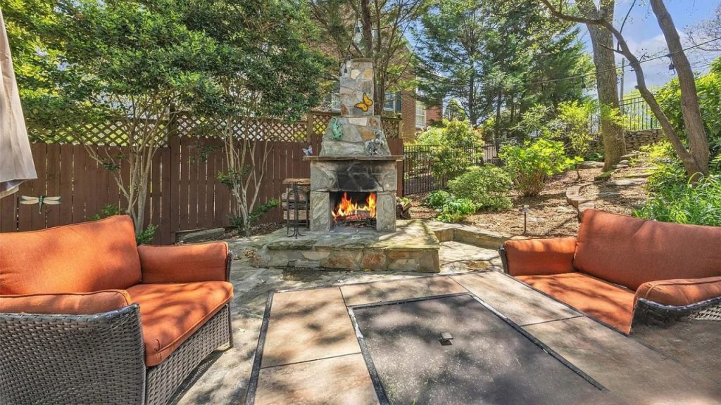 A scenic outdoor patio with a stone fireplace, orange wicker chairs, and lush greenery under a sunny sky.