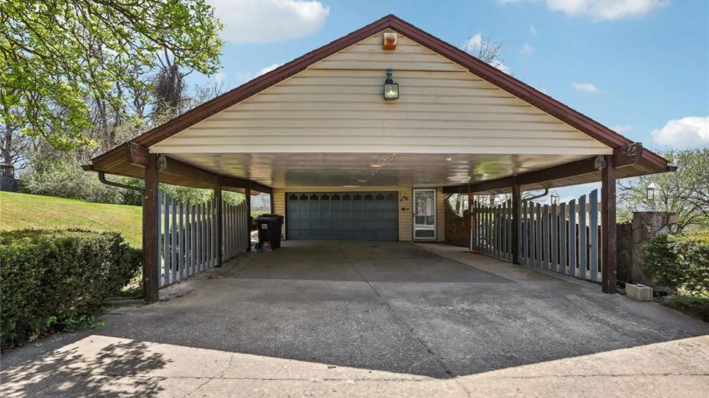 A spacious carport with wooden beams, beige siding, and a decorative garage door, surrounded by greenery under a bright sky.